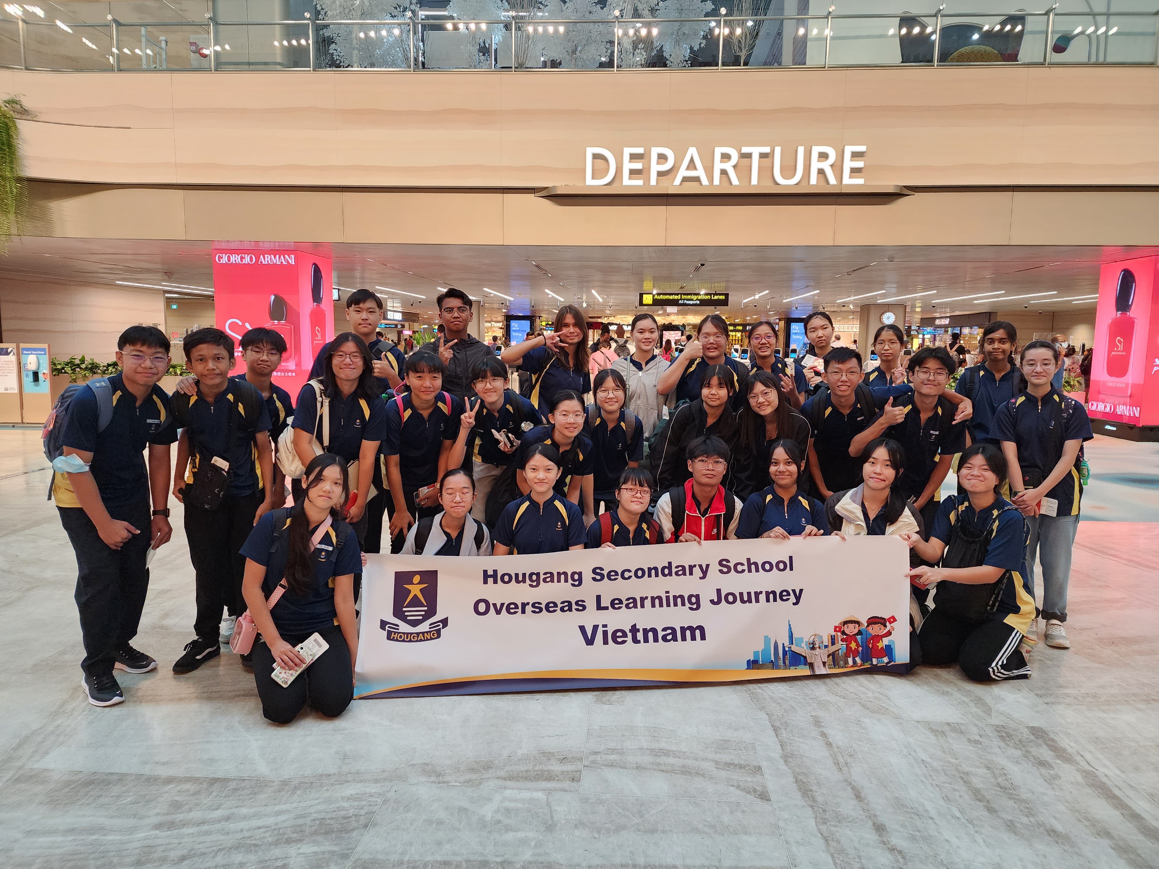 Students in navy uniforms pose at an airport departure hall holding a Hougang Secondary School Vietnam Learning Journey banner.
